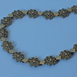 May include: A gold-toned necklace featuring an ornate design. The necklace is composed of repeating floral and diamond-shaped links. The intricate details and warm color suggest a vintage or antique style, suitable for special occasions or adding a touch of elegance.
