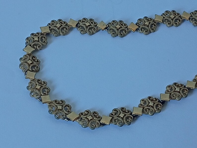 May include: A gold-toned necklace featuring an ornate design. The necklace is composed of repeating floral and diamond-shaped links. The intricate details and warm color suggest a vintage or antique style, suitable for special occasions or adding a touch of elegance.