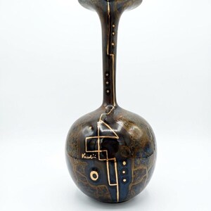 Può includere: Un vaso in ceramica marrone scuro e blu con finitura lucida, caratterizzato da un collo lungo e sottile e un corpo arrotondato. La superficie è decorata con linee geometriche dorate, punti e la firma Vusali, creando un motivo distintivo.
