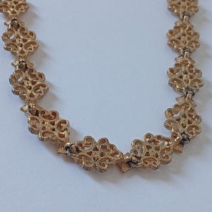 May include: A gold-colored necklace with an ornate, openwork design. The necklace features repeating floral or scroll-like patterns, connected by small links. The intricate details and warm metallic tone suggest a vintage or antique aesthetic.