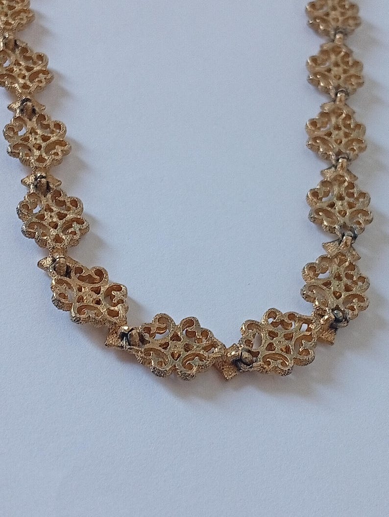May include: A gold-colored necklace with an ornate, openwork design. The necklace features repeating floral or scroll-like patterns, connected by small links. The intricate details and warm metallic tone suggest a vintage or antique aesthetic.