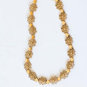 May include: A gold-toned necklace with an ornate design. The necklace features repeating floral-like patterns and diamond-shaped accents. The chain is connected with a clasp. The jewelry is displayed on a white background.