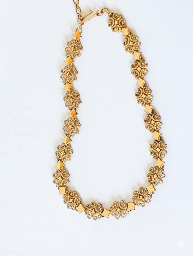 May include: A gold-toned necklace with an ornate design. The necklace features repeating floral-like patterns and diamond-shaped accents. The chain is connected with a clasp. The jewelry is displayed on a white background.