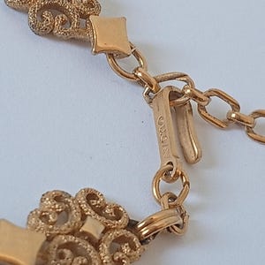 May include: Close-up of a gold-toned necklace with ornate filigree detailing. The necklace features a decorative clasp and chain links. The brand name "AVON" is visible on the clasp.