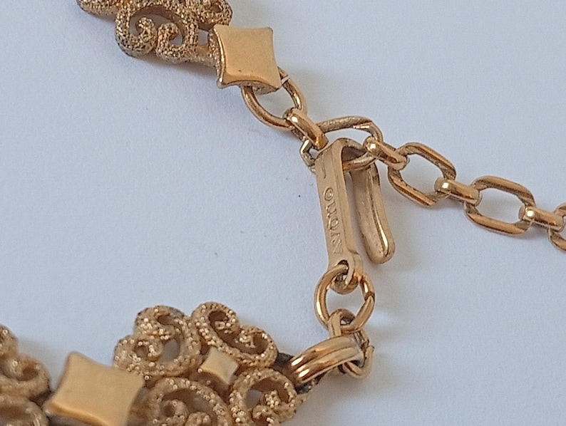May include: Close-up of a gold-toned necklace with ornate filigree detailing. The necklace features a decorative clasp and chain links. The brand name "AVON" is visible on the clasp.
