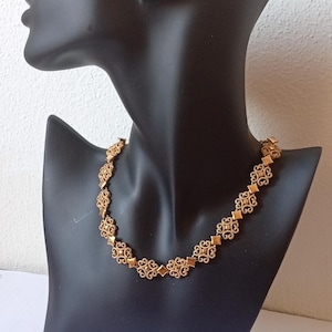May include: A gold-colored necklace with an ornate, repeating pattern of diamond and floral shapes. The necklace is displayed on a black mannequin bust against a white background. The necklace is a statement piece.