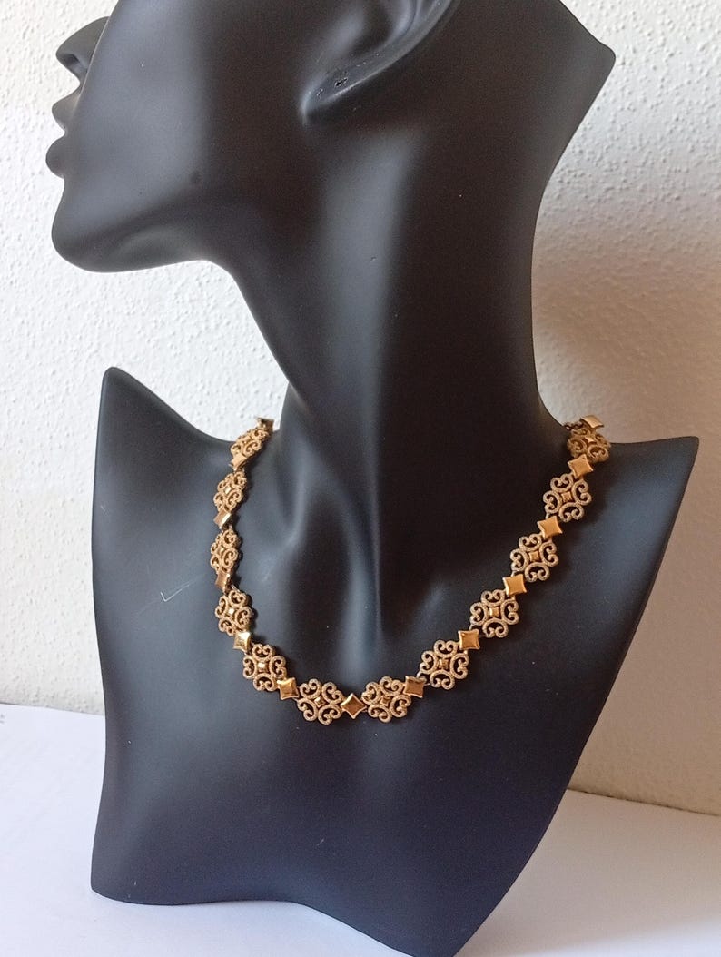 May include: A gold-colored necklace with an ornate, repeating pattern of diamond and floral shapes. The necklace is displayed on a black mannequin bust against a white background. The necklace is a statement piece.