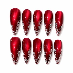 May include: Set of ten glossy, deep red, almond-shaped false nails. Each nail features a silver, swirling design with small, silver bead accents near the tip. The nails are arranged on a white background, showcasing their vibrant color and intricate details.