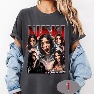 Könnte beinhalten: Ein anthrazitfarbenes T-Shirt mit einem Grafikdruck, der oben den Text NIKKI in fetten roten Buchstaben und unten Obsession in Schreibschrift zeigt. Das Design umfasst fünf Porträts einer Person, umgeben von weißen Blitzen auf rotem Grund.