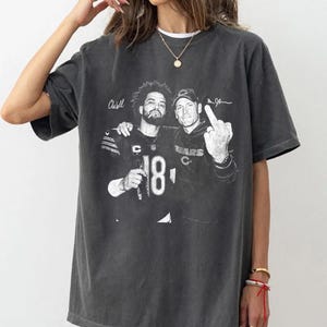 May include: A charcoal gray short-sleeve t-shirt featuring a black and white graphic print of two football players. One player wears a jersey with the number 18 and the other wears a cap. The text "CWill" and "Ben Johnson" is printed above them.