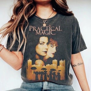 Könnte beinhalten: Ein anthrazitfarbenes Kurzarm-T-Shirt mit einem Grafikdruck von zwei Frauengesichtern über einer Reihe leuchtender gelber Kerzen und Silhouetten von Figuren, die Regenschirme halten. Der Text "PRACTICAL MAGIC" ist in einer Serifenschrift darüber gedruckt.
