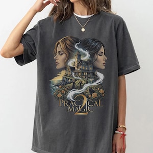 May include: A charcoal grey short-sleeve t-shirt featuring a graphic print of two women in profile facing a large house on a cliffside. Below the house are yellow roses and the text "PRACTICAL MAGIC 2" in a gold serif font with swirling white smoke.