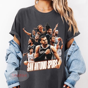 Pode incluir: Uma t-shirt de manga curta cinzento-carvão com uma colagem gráfica de vários jogadores de basquetebol com uniformes dos San Antonio Spurs. O texto "SAN ANTONIO SPURS" está impresso numa fonte a negrito e clara na parte inferior do design.