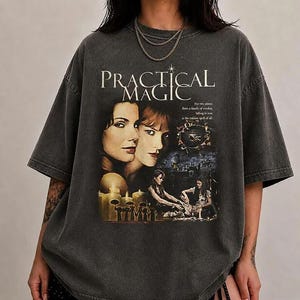 Könnte beinhalten: Ein anthrazitfarbenes Oversize-T-Shirt mit einem Grafikdruck zum Film Practical Magic. Das Design zeigt Porträts von zwei Frauen, Text sowie Szenen mit Kerzen und Figuren. Das T-Shirt hat einen Rundhalsausschnitt und kurze Ärmel.