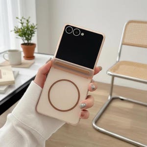 May include: A light pink phone case with a circular design and a black screen. The phone case is held in a hand. The background includes a small potted plant, a mug, and a chair.