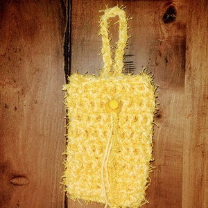 May include: A yellow crocheted bag with a yellow button and a long fringe. The bag is hanging on a wooden surface.