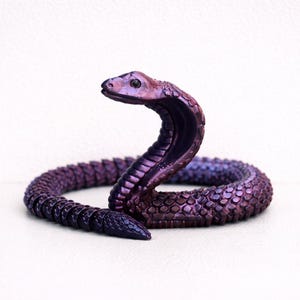 May include: A purple cobra ornament with a detailed scale pattern. The snake is coiled, with its head raised. The color transitions from a deep purple to a lighter hue, creating a gradient effect. The ornament is set against a white background.