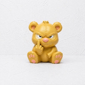 May include: A yellow figurine of an angry bear with a pink nose and paws, giving the middle finger. The bear has a furrowed brow and is sitting down. The background is a plain white surface.