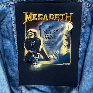May include: A black patch with the gold band name "MEGADETH" and an illustration of a skeleton kneeling before a tombstone reading "RIP MARY JANE". The patch is attached to a denim jacket.