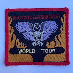 Peut inclure: Un écusson brodé carré à bordure rouge portant le texte BLACK SABBATH en rouge et WORLD TOUR en jaune. Le motif central représente un crâne ailé avec des cornes dans des tons de violet et de gris, surmonté d'un petit cercle contenant 666.