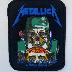 Puede incluir: Parche cuadrado negro con el nombre de la banda "METALLICA" en azul, sobre una calavera con un sombrero verde. La calavera tiene un gráfico de cerebro debajo, con el texto "CRASH COURSE IN BRAIN SURGERY" en azul.