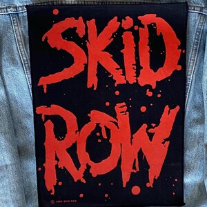 May include: A black patch featuring the words "SKID ROW" in large, red, dripping letters. The patch is attached to a light blue denim jacket. Small red splatters and the text "© 1989 SKID ROW" are also visible.