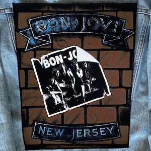May include: A light blue denim jacket with a large Bon Jovi patch. The patch shows a brick wall design, the band's name in a banner, and the words "New Jersey" below. A black and white image of the band is also on the patch.