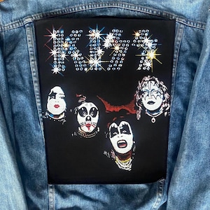 May include: A light blue denim jacket with a large black patch featuring the band KISS. The patch displays the band's name in a sparkling, star-like font above four band member portraits in makeup.