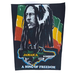Op de afbeelding: Een zwarte stoffen patch met een afbeelding van Bob Marley met dreadlocks voor een rood, goud en groene vlag. Daaronder staat een kaart van Jamaica met de tekst JAMAICA, KINGSTON en de zin A SONG OF FREEDOM in het wit gedrukt.