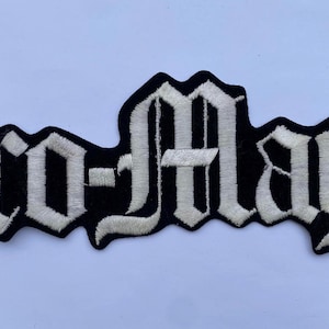 May include: A black embroidered patch featuring the text Cro-Mags in a white gothic-style font. The patch has a die-cut shape that follows the contours of the lettering, with a thick black border surrounding the white stitched characters.