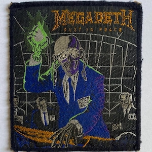 May include: Embroidered patch featuring the Megadeth band logo and the words "Rust in Peace." The patch depicts a skeletal figure in a blue suit, holding a green object, with a black background and additional figures.