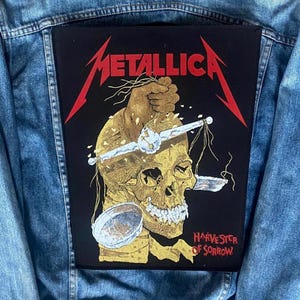 May include: A denim jacket with a large, black patch featuring the band name "Metallica" in red, stylized lettering. The patch depicts a gold skull with a hand holding a scale, and the words "Harvester of Sorrow."