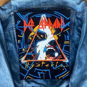 Könnte beinhalten: Eine blaue Jeansjacke mit einem großen rechteckigen Aufnäher auf dem Rücken. Der Aufnäher zeigt ein farbenfrohes Design mit dem Bandnamen "Def Leppard" und dem Wort "Hysteria". Das Design umfasst ein Dreieck und geometrische Formen.