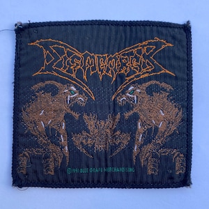 May include: A square black fabric patch featuring an orange embroidered logo that reads DISMEMBER at the top. Below the text are two brown, snarling, creature-like figures facing each other. The text 1991 BLUE GRAPE MERCHANDISING is printed in green.