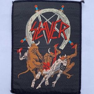 May include: A rectangular black fabric patch featuring an embroidered Slayer logo in red letters above a white pentagram and sword design. Below, three brown horned demon figures are depicted in a dynamic scene with a pale, skeletal figure.