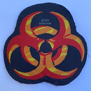 May include: A black embroidered patch featuring a red and yellow biohazard symbol. The design includes the text "(c) 1994 BIOHAZARD" printed in small letters above the central circular element of the symbol. The patch has a dark fabric base.