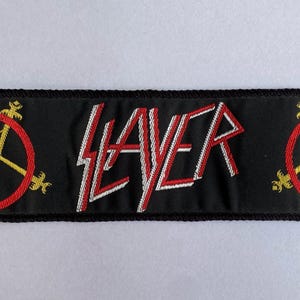 May include: Black embroidered patch with the word "SLAYER" in red and white lettering. The patch features a red circle with a yellow star and decorative elements on each end.