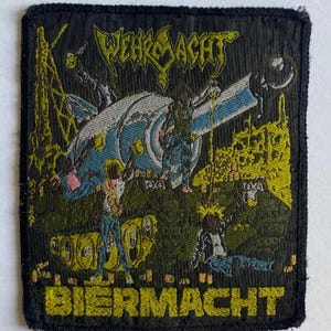Könnte beinhalten: Schwarzer, gestickter Aufnäher mit den gelben Worten "WEHRMACHT" und "BIERMACHT". Der Aufnäher zeigt eine Cartoon-Illustration einer futuristischen Szene mit Figuren und Maschinen in Blau, Gelb und Rosa.