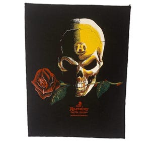 May include: A black rectangular fabric patch featuring a yellow and white skull graphic with a letter A on the forehead. A red rose is held in the skull's teeth. Below the skull, the text reads Alchemy battle-dress in red and white.