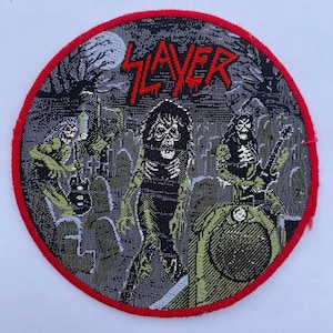 Op de afbeelding: Een ronde geweven patch met een rood Slayer-logo aan de bovenzijde. De afbeelding toont drie skeletachtige figuren op een begraafplaats met een volle maan op de achtergrond. De patch is afgewerkt met een felrode gestikte rand.