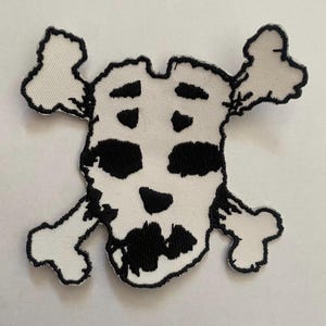 May include: A white embroidered patch featuring a black skull and crossbones design. The skull has stylized black eye sockets, a nose, and a mouth, with two crossed bones extending from behind the head. The edges are finished with black stitching.