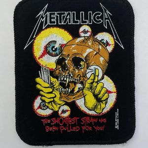 Peut inclure: Écusson noir avec le mot "METALLICA" en blanc, et un motif de crâne et d'œil. Le crâne est marron et les yeux sont jaunes et rouges. Le texte "THE SHORTEST STRAW HAS BEEN PULLED FOR YOU!" est en rouge.