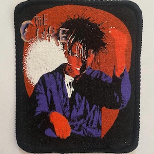 May include: A rectangular fabric patch with a black border featuring a graphic of a person with dark, messy hair wearing a purple jacket. The background includes a large orange circle and the text THE CURE in a stylized, light-colored font.