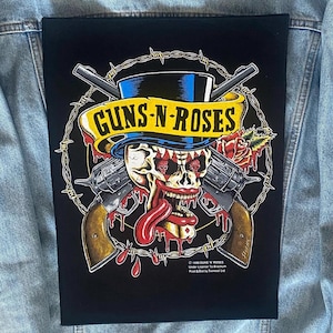 May include: Black patch featuring a Guns N' Roses design. The graphic includes a skull wearing a top hat, crossed revolvers, a banner with the band's name, and a rose. The design is surrounded by barbed wire.
