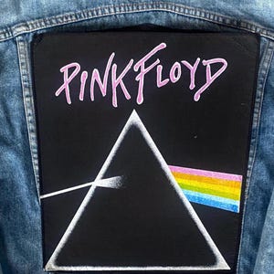 Może przedstawiać: Kurtka dżinsowa z dużą, prostokątną naszywką z napisem "PINK FLOYD" w różowym kolorze. Poniżej biały trójkąt z tęczowym spektrum.