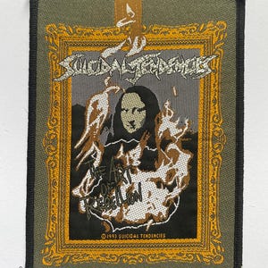 Peut inclure: Écusson tissé avec l'inscription "Suicidal Tendencies" en argenté délavé au-dessus d'une image d'un visage de femme entouré de flammes. Le texte "The Art of Rebellion" est en dessous. L'écusson est encadré en or.