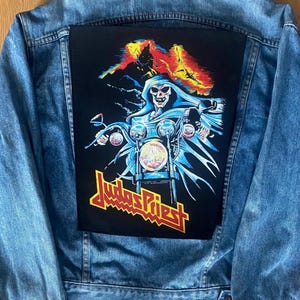 Pode incluir: Casaco de ganga azul claro com um grande patch preto nas costas. O patch apresenta um esqueleto a andar de mota com o nome da banda "Judas Priest" em amarelo e vermelho.