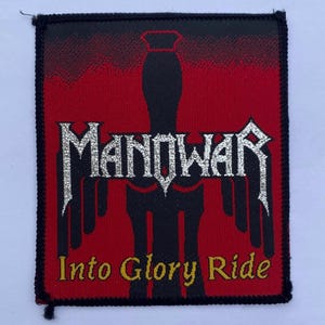 Puede incluir: Un parche bordado rectangular que presenta la silueta de una espada negra centrada sobre un fondo rojo. La palabra MANOWAR está bordada en hilo metálico plateado sobre el texto amarillo Into Glory Ride, todo rodeado por un borde negro.