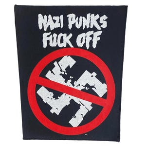 May include: A black rectangular fabric patch featuring the white text NAZI PUNKS FUCK OFF at the top. Below the text is a white swastika symbol crossed out by a bold red circle and a diagonal red line across the center of the design.