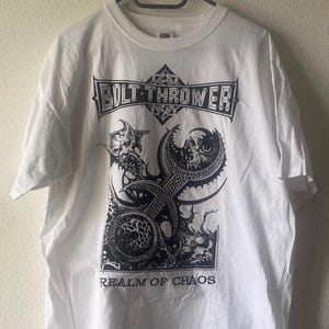 Puede incluir: Camiseta blanca con un diseño gráfico negro. El diseño incluye el texto "BOLT THROWER" y "REALM OF CHAOS", junto con una calavera y tentáculos. La camiseta está colgada en una percha negra.